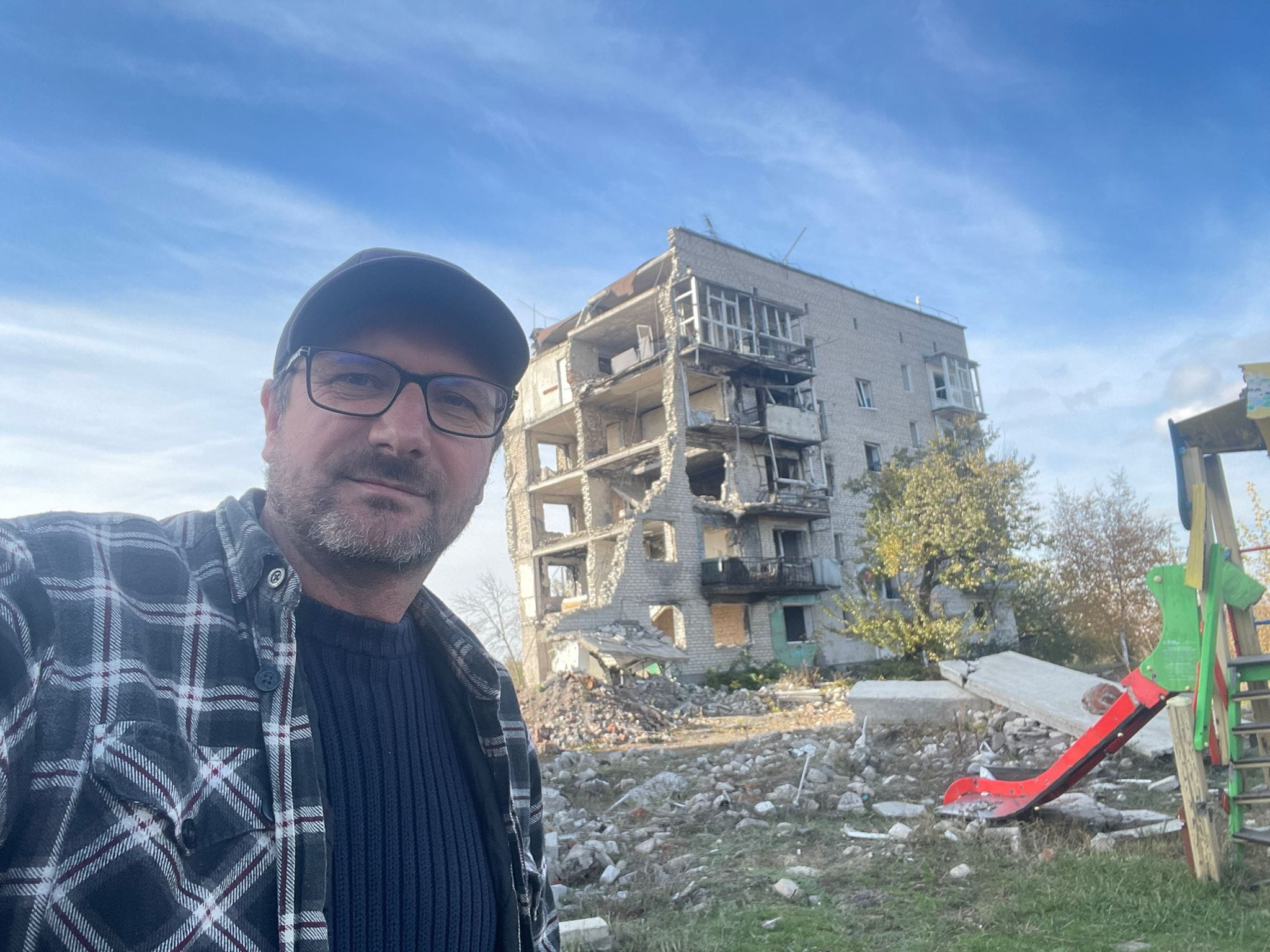 Csaba wearing checked shirt and baseball cap, standing in front of a bombed-out apartment block in Ukraine, blue sky behind